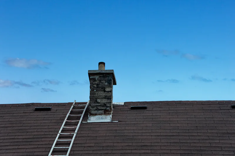 Ladder leaned on a dark roof beside a brick chimney