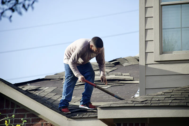 Roofer using a tear-off shovel to strip old shingles beside a second-story wall